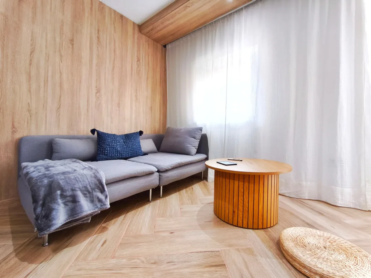 Rincón de estar: sofá gris con cojines, mesa baja redonda de madera y una cortina translúcida que filtra la luz de la terraza.